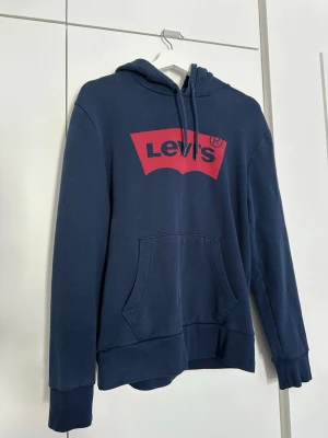 Blå Levi's hoodie med röd logga - Mörkblå hoodie från Levi's med stor röd logga på bröstet. Klassisk känguruficka framtill, huva med dragsko och ribbade muddar vid ärmar och nederkant. Storlek M. 