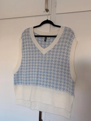 Ljusblå och vit stickad väst - Trendig stickad väst från H&M i ljusblått och vitt med klassiskt rutigt hundtandsmönster. Västen har v-ringning och breda ribbade kanter. Perfekt att styla över en skjorta eller t-shirt för en cool lager-på-lager-look. Obs. hund finns i hemmet