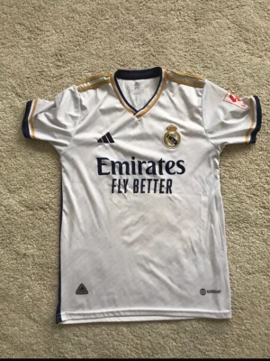 Real Madrid matchtröja Vini Jr. - Säljer en vit Real Madrid matchtröja från Adidas med Vini Jr. och nummer 7 på ryggen. Tröjan har gulddetaljer på axlar och ärmslut, klubbmärke och sponsortryck framtill samt marinblå inslag. Perfekt för fotbollsfans!
