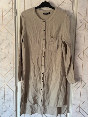 Beige långärmad skjortklänning KappAhl - Stilren beige skjortklänning från KappAhl med lång ärm, rund halsringning och knappar hela vägen framtill. Klänningen har en bröstficka. Artikelnummer 72923/201103