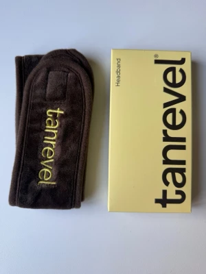 Brunt mjukt pannband hårband headband från Tanrevel - Säljer ett brunt headband från Tanrevel i mjukt,  material med broderad logga i gult. Perfekt för att hålla håret borta vid sminkning, hudvård eller vid brun utan sol. Snygg och enkel design med kardborrestängning för enkel användning. Helt oanvänt. Omkrets på ca 58 cm och tillverkat av mjuk velour.