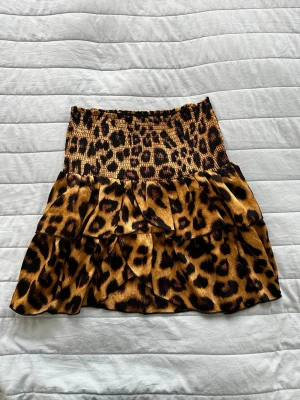 Leopardmönstrad volangkjol Neo Noir - Kort kjol från Neo Noir med leopardmönster i bruna och svarta toner. Kjolen har bred smock i midjan och flera lager volanger som ger en snygg och trendig look. Perfekt för dig som vill sticka ut med ett djurmönster. Storlek S, stretching material så den passar även M. Nyskick