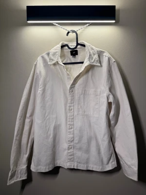 Vit skjorta i bomull från H&M - Klassisk vit skjorta från H&M i mjuk bomull med rak passform. Skjortan har lång ärm, enkel bröstficka och knappar framtill. Perfekt basplagg med stilren look och normal krage.