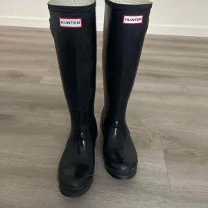 Svarta stövlar från Hunter - Snygga svarta stövlar från Hunter, perfekta för regniga dagar. De har en klassisk design med justerbart spänne på sidan och Hunter-loggan framtill. Insidan har ett mönstrat foder för extra komfort.