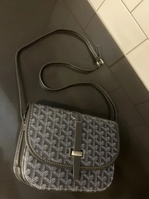 axelväska - Snygg axelväska i svart och grått med grafiskt mönster och lång axelrem. Väskan har lock med spänne framtill och detaljer i konstläder. Skickas inom 24 timmar färgen är lite ljusare i värkligheten