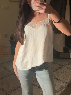 Vit linnetopp med spetsdetaljer - Snygg vit linnetopp och v-ringad halsringning dekorerad med spets. Toppen har en lös passform och är perfekt för varma dagar. Storlek XS