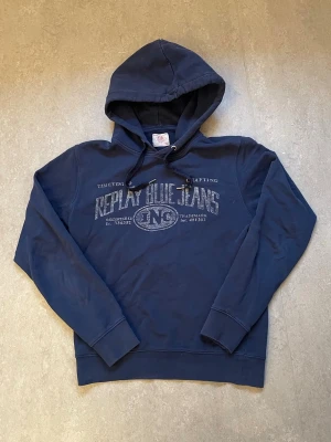 Marinblå hoodie Replay Blue Jeans - Snygg marinblå hoodie från Replay Blue Jeans med tryck framtill i vitt och silver. Klassisk huva med dragsko och ribbade muddar vid ärmar och nederkant. Perfekt för en avslappnad streetwear-look. Mjuk bomullsblandning för skön känsla.