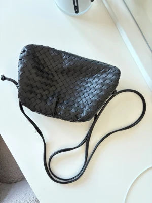 Svart flätad axelväska - Snygg svart handväska med flätad design och lång axelrem. Perfekt för dig som gillar trendiga accessoarer med unik form och stilren look. En liten fläck inuti väskan som syns på fjärde bilden. 