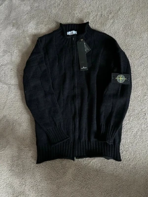 Svart stickad ziptröja Stone Island - Snygg svart stickad ziptröja från Stone Island med ribbade muddar och hög krage. Tröjan har dragkedja framtill och den klassiska Stone Island-loggan på vänster ärm. Perfekt för lager-på-lager och streetwear-stil.