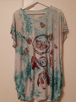 Mönstrad topp med fjärilar och drömfångare - Unik topp med korta ärmar och rund hals. Tryck med drömfångare, fjädrar och orange fjärilar på en turkos och vit bakgrund. Materialet känns mjukt och lätt, perfekt för en avslappnad stil. Glittriga detaljer ger extra wow-faktor.
