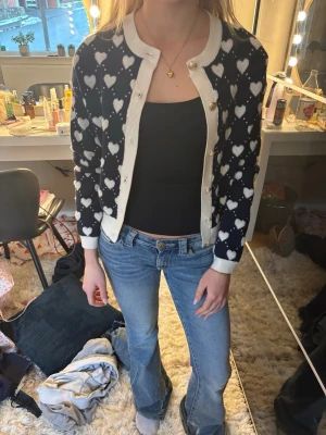 Mörkblå kofta med hjärtmönster - Supersöt mörkblå kofta från Holly & White med vita hjärtan över hela och kontrasterande vita kanter. Rund halsringning och guldfärgade knappar framtill. Perfekt för att piffa upp en enkel outfit.