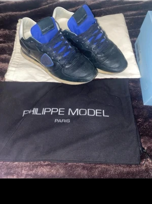 Philippe Model svarta sneakers i skinn - Snygga svarta sneakers från Philippe Model med blåa detaljer och vit insida. Skorna har klassisk snörning, rund tå och en beige sula. Tillverkade i skinn med textil på plösen och sidan. Perfekta för dig som gillar stilrena men ändå unika sneakers. Skorna är även knappast använda, köpte de i början av november sedan har jag använt de ett väldigt få par gånger. Du får även med Lanvin dustbag och deras egna dustbag samt box. För ytterligare bilder och funderingar får du gärna höra av dig. (: