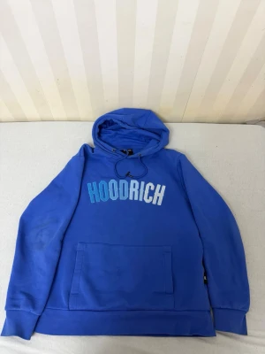Blå HOODRICH hoodie med tryck - Snygg blå hoodie från HOODRICH med stor magficka och huva med dragsko. Framtill står det HOODRICH i vitt och blått tryck. Hoodien har ribbade muddar vid ärmar och nederkant. Perfekt för en chill och street-inspirerad look.