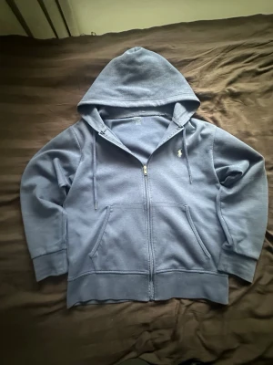 Blå zip hoodie från Polo Ralph Lauren - Mörkblå hoodie med dragkedja från Polo Ralph Lauren. Klassisk design med huva, dragsnören och två stora fickor framtill. Ikoniska vita loggan broderad på bröstet. Tillverkad i mjukt bomullsmaterial som är skönt att ha på sig.