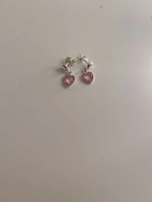 Söta örhängen med rosa hjärtan - Ett par silverfärgade örhängen med små hängande hjärtan dekorerade med rosa stenar. Hjärtana har en öppen design och örhängena har klassisk stiftknäppning. Perfekta för dig som gillar gulliga accessoarer och vill addera lite färg till din look.
