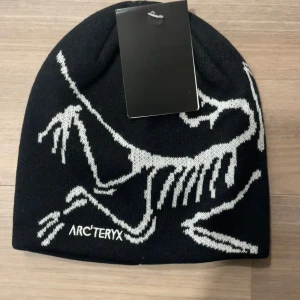 Svart mössa från Arc'teryx - Snygg svart mössa från Arc'teryx med vit stickad dinosaurie-design och logga framtill. Klassisk passform och mjukt stickat material som håller dig varm. Perfekt accessoar för kalla dagar och streetstyle.