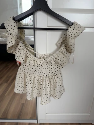 Prickig volangtopp med snörning - Supersöt vit topp med svarta prickar och volangdetaljer. Toppen har snörning i ryggen och breda axelband med volanger. Perfekt för dig som gillar romantisk och lekfull stil. Tillverkad i ett lätt och luftigt material.