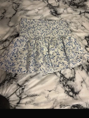 Kjol - Supersöt kortkjol från H&M i vitt med blått blommönster. Kjolen har smockad midja som ger en bekväm passform och volang nedtill för en extra feminin touch. Perfekt för varma dagar och enkel att matcha med en enkel topp.