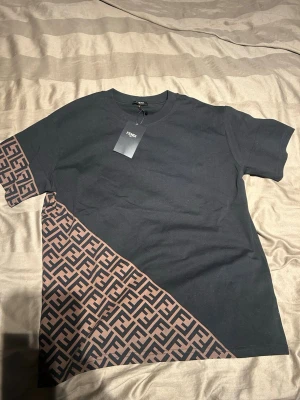 Svart Fendi t-shirt  - Snygg svart t-shirt från Fendi med diagonal brun logomönstrad detalj över ena ärmen och nedre delen av tröjan. Klassisk rund halsringning och korta ärmar. Perfekt för dig som vill sticka ut med en exklusiv streetwear-look.