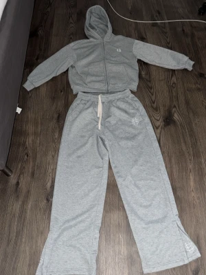 Grått loungewear-set med huva - Mysigt grått loungewear-set med zip-hoodie och vida byxor. Hoodien har dragkedja, fickor och en broderad logga på bröstet. Byxorna har resår i midjan med vit dragsko och matchande logga på benet. Perfekt för chill dagar hemma.