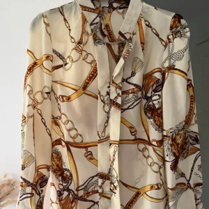 Beige mönstrad skjorta med kedjor - Snygg beige skjorta med unikt kedjemönster i guld och brunt. Skjortan har klassisk krage och långa ärmar, tillverkad i ett lätt och glansigt material som ger en lyxig känsla. 