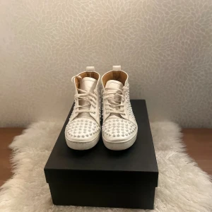 Vita Louboutin sneakers med nitar - Säljer ett par vita high-top sneakers från Christian Louboutin med ikoniska röda sulor och coola nitar över hela ovandelen. Skorna har snörning och är tillverkade i skinn, vilket ger en lyxig känsla. Perfekt för dig som vill sticka ut med din stil. Pris går att diskutera 