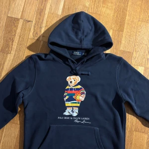 Mörkblå Polo Bear hoodie från Ralph Lauren - Säljer en mörkblå hoodie från Polo Ralph Lauren med ikoniska Polo Bear-trycket på bröstet. Tröjan har huva med snörning och känguruficka. Björnen på framsidan har en färgglad randig tröja och håller en basketboll. Perfekt statement-plagg för dig som gillar streetstyle.
