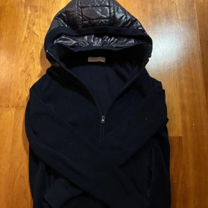 Blå sayless cardigan - Blå cardigan jacka från sayless med avtagbar quiltad huva i glansigt material. Jackan har dragkedja framtill och två sidofickor med dragkedja. Stickad struktur i kroppen och ribbade muddar vid ärmslut och nederkant. Perfekt för dig som gillar stilren och funktionell design.