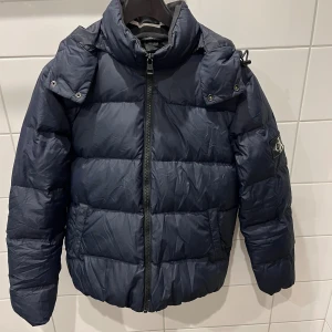 Mörkblå pufferjacka Calvin Klein Jeans - Säljer en mörkblå pufferjacka från Calvin Klein med huva och dragkedja framtill. Jackan har broderad logga på ärmen och tryckt logga i nacken. Perfekt för kalla dagar och har en riktigt schysst streetstil.