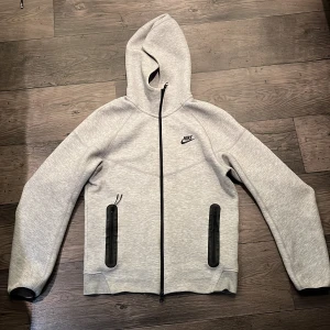 Nike tech hoodie - Nike hoodie i ljusgrått med svart logga på bröstet. Tröjan har hel dragkedja framtill, två svarta dragkedjeförsedda fickor och huva. Perfekt för chill eller träning, med en clean och sportig look.