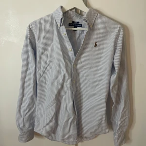Randig skjorta från Ralph Lauren - Klassisk slim fit skjorta från Ralph Lauren i ljusblått och vitt med smala ränder. Skjortan har lång ärm, knappar framtill och ikonisk broderad logga på bröstet. I mycket fint skick. Öppen för pris förslag