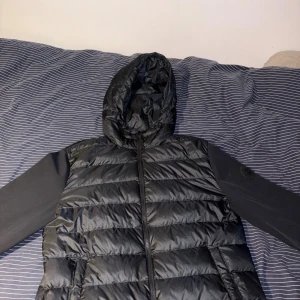 Svart pufferjacka från GANT med huva - Svart pufferjacka från GANT med huva och dragkedja framtill. Jackan har quiltad kropp och släta ärmar med GANT-logga på vänster ärm. Perfekt för kyliga dagar och har en clean, sportig look.