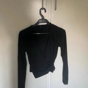Svart omlottopp med knyt - Säljer en svart omlottopp med lång ärm och snygg knytning framtill. Toppen har en djup v-ringning och är croppad i modellen, vilket ger en trendig look. Materialet är ribbat och känns mjukt mot huden. Perfekt att styla med höga jeans eller kjol. Slutsåld men supertrendig!!