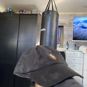 Svart keps från Polo Ralph Lauren - Svart keps från Polo Ralph Lauren med klassisk broderad logga framtill och röd brodyr bak. Justerbart spänne i brunt skinn baktill. Snygg och tidlös modell i bomull, perfekt för att lyfta din outfit.