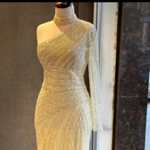 Glittrig champagnefärgad långklänning - Supersnygg långklänning i champagnefärg med glittriga detaljer och ett unikt oneshoulder-snitt. Klänningen har en hög halsringning och en lång ärm, medan andra axeln är bar. Materialet är glittrigt och skimrande, perfekt för att sticka ut på bal eller fest.