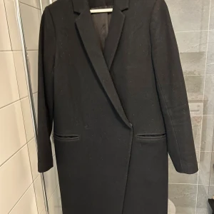 Svart kappa från Wera Stockholm - Stilren svart kappa från Wera Stockholm i klassisk rak modell med snygg krage och diskreta fickor. Kappan är helsvart och har långa ärmar, perfekt för dig som gillar minimalistisk och tidlös design. Materialet är mjukt och känns lyxigt.