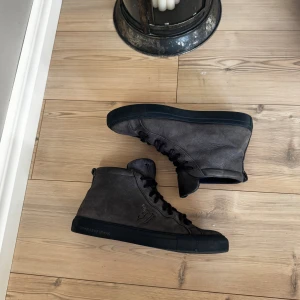 Blåa höga sneakers från Trussardi Jeans - Säljer ett par snygga höga sneakers från Trussardi Jeans i mörkblått skinn. Skorna har svart sula, snörning och logga på sidan. Stilren design med rund tå och mjuk insida för extra komfort. Perfekta för dig som gillar streetstyle och vill sticka ut.