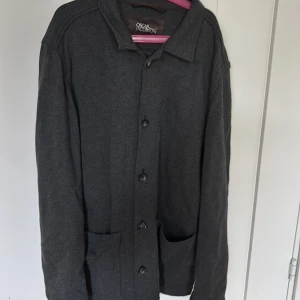 Mörkgrå overshirt från Oscar Jacobson - Stilren mörkgrå overshirt från Oscar Jacobson med klassisk krage, knäppning framtill och två stora fickor. Perfekt lager-på-lager-plagg med avslappnad passform och mjukt material. Enkel att matcha till olika outfits.