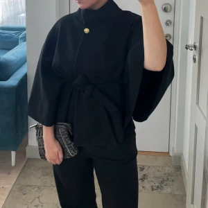 Svart poncho från missay  - Stilren svart poncho med knytbälte i midjan och vida, trekvartsärmar. Ponchon har hög krage och en guldig knapp upptill. Perfekt för dig som gillar minimalistisk och trendig look.