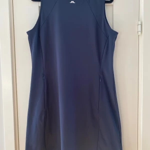 Marinblå ärmlös golfklänning J.Lindeberg - Snygg marinblå klänning från J.Lindeberg med vit krage och diskret logga framtill. Modellen är ärmlös och har en sportig, slim passform med dragkedja i sidan. Materialet är mjukt och stretchigt polyester som ger en skön känsla. Perfekt för dig som gillar stilren design. Mått Över bröstet: ca 53 cm Över höfterna: ca 51 cm Längd från axeln och ner: ca 90 cm  79% polyester och 21% elastan   Kommer från ett djur och rökfritt hem.