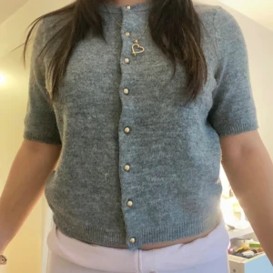 Grå stickad kofta från H&M - Mysig grå stickad kofta från H&M med korta ärmar och snygga guldiga knappar framtill. Perfekt att slänga på sig över ett linne eller t-shirt.