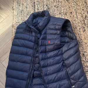 Mörkblå dunväst från Polo Ralph Lauren - Snygg mörkblå dunväst från Polo Ralph Lauren med quiltad design och dragkedja framtill. Västen har två sidofickor med dragkedja och klassisk ståkrage. Den ikoniska röda Polo-loggan är broderad på bröstet. Perfekt för lager-på-lager under kyliga dagar.