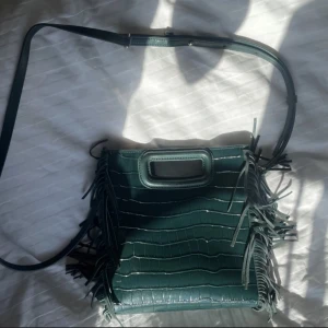 Grön axelväska med fransar - HEJ!nu säljer jag min älskade och snygga mörkgrön lilla Maje väska med krokodilpräglat mönster och fransar längs sidorna.Denna bild är lånad då jag ej är hemma,bara o skriva privat om bilder.perfekt för att ge din outfit en cool touch.Den e använd runt 10 gånger och säljs för den ej kommer till användning,skriv för fler bilder❤️