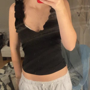 Nelly linne - Säljer en svart croppad linnetopp med breda axelband och volangdetaljer längs kanterna. Toppen har en djup v-ringning och sitter tight mot kroppen. Perfekt för dig som gillar en stilren men ändå söt look.