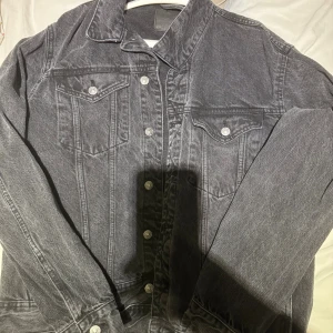 Svart jeansjacka med silvriga knappar - Snygg svart jeansjacka med klassisk krage och silvriga knappar framtill. Jackan har två bröstfickor med lock och knapp samt raka sömmar för en stilren look. Perfekt för dig som gillar en tidlös och cool stil.