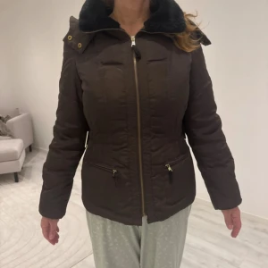 Brun pufferjacka med huva - Snygg brun pufferjacka med dragkedja framtill, två fickor med guldfärgade detaljer och en mysig svart krage. Jackan har huva och långärmad design, perfekt för kalla dagar. Passformen är normal och materialet känns som polyester.