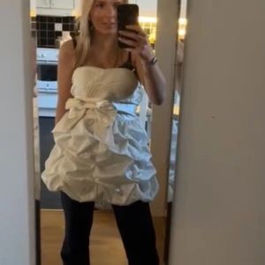 Vit kort klänning med rosett - Säljer en vit kort klänning med axelbandslös design och stor rosett framtill. Klänningen har volangdetaljer och ett snyggt rynkat bakstycke. Materialet är glansigt och känns lyxigt, perfekt för dig som vill sticka ut med en unik look. Perfekt för studenten