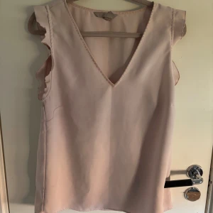 Ljusrosa blus med volangärmar - Säljer en ljusrosa blus med v-ringning och volangdetaljer vid ärmsluten. Blusen är ärmlös och har en mjuk, flowy passform som känns skön mot huden. Perfekt för dig som gillar en feminin och enkel stil.