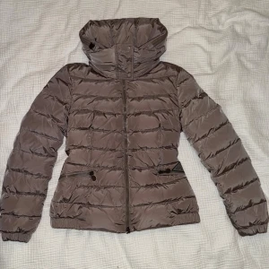 Moncler brun dunjacka, storlek 2 - Brun Moncler dunjacka med huva. Jackan har dragkedja och fickor med dragkedja fram. Den har en figurnära passform med resar i midjan bak. Mycket fint skick. Storlek 2 Självklart 100% äkta