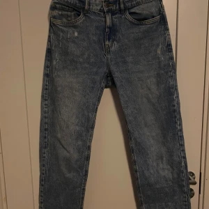 Blå jeans från Lindex, storlek 170 - Säljer ett par klassiska blå jeans från Lindex i storlek 170. Jeansen har en rak passform med fem fickor och slitna detaljer framtill för en cool vintagekänsla. Materialet är robust denim med ljus tvätt och kontrastsömmar. Perfekt för dig som gillar avslappnad stil.